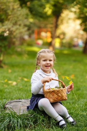 Smiling young blonde girl sitting in the grass collecting leaves with basket