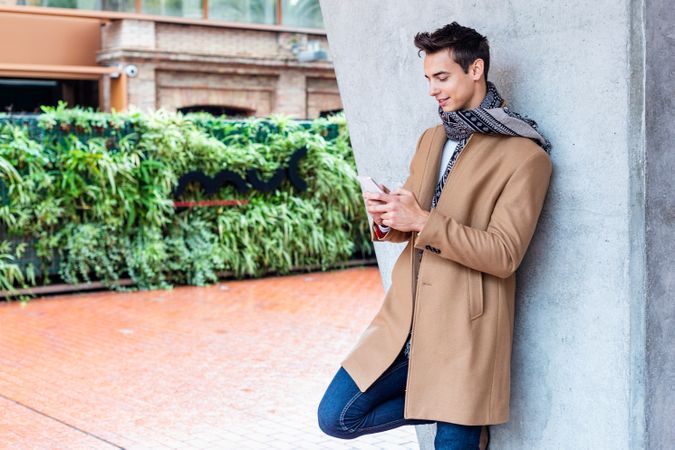 Man in camel coat and scarf looking at phone while leaning on outdoor wall