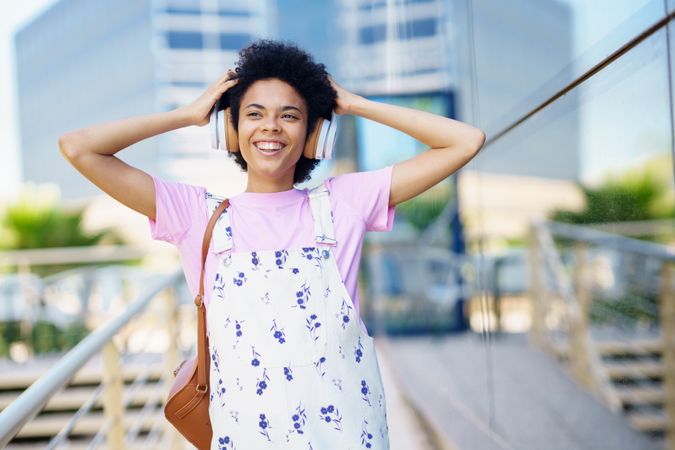 Joyful female in floral overalls walking outside listening to headphones touching hair