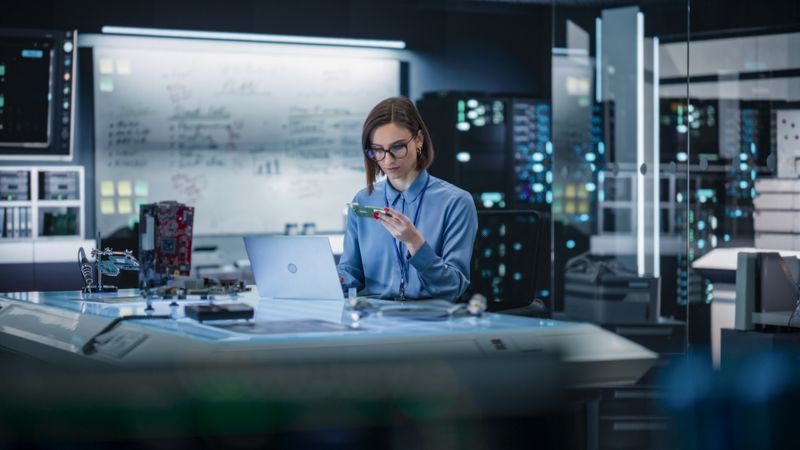 Portrait of Female Specialist Using Laptop Computer to Improve Electronic Circuit Board in Modern Office. Professional Engineer Thinking and Typing Ideas for Artificial Intelligence Project