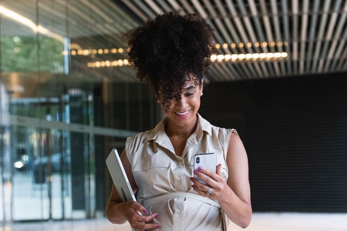 Smiling Black pregnant woman using smartphone and carrying laptop