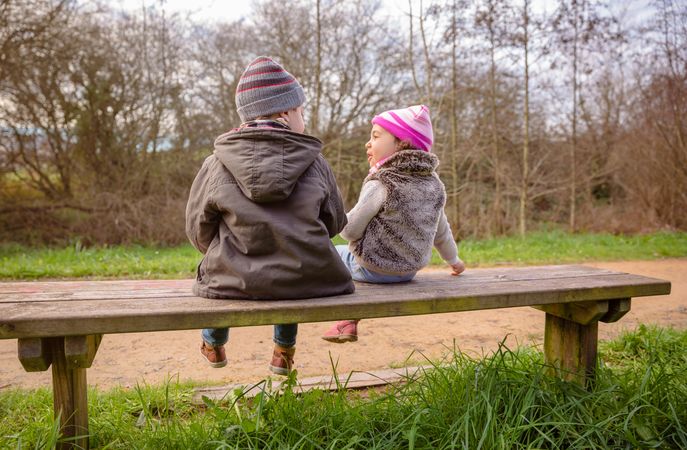 Back of little boy and girl sitting on park bench chatting in forest