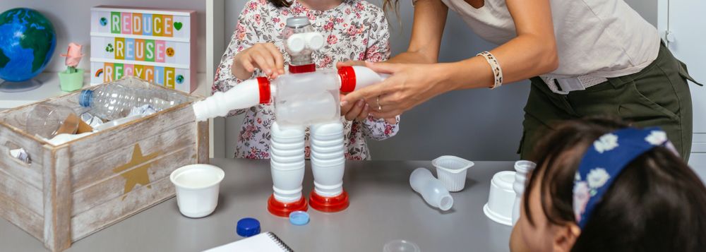 Teacher helping to girl make recycled toy robot with plastic packages