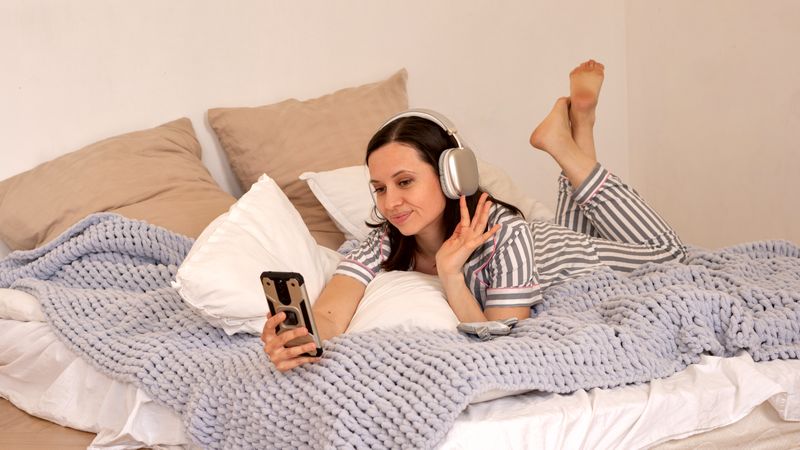 Woman in bed listening to music on smartphone, waving hand.