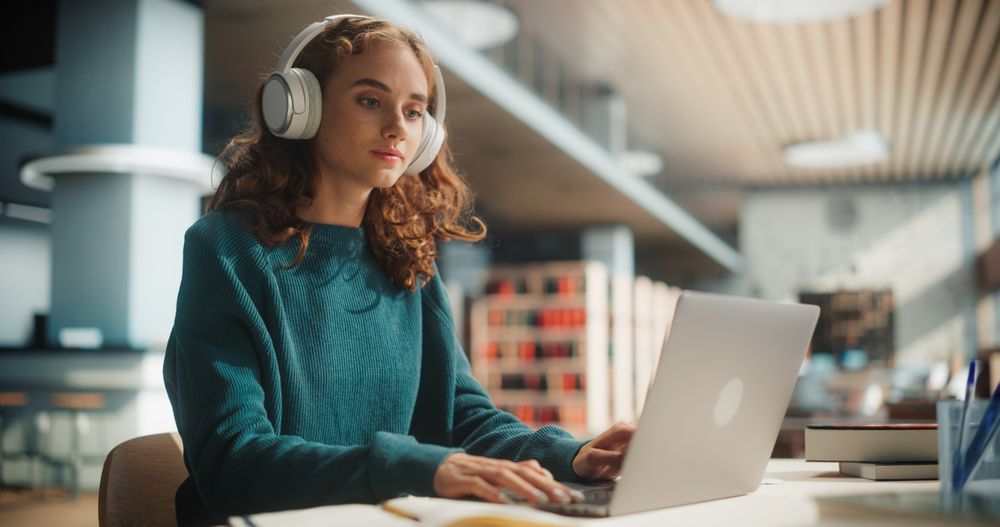 Concentrated Young Female Student With Headphones Using Laptop In Modern Library.