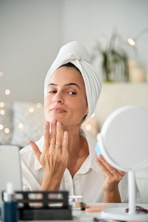 Attractive female in towel turban applying facial cream at mirro