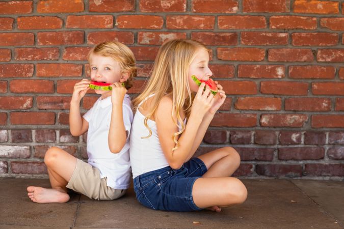 Cute Young Boy and Girl Eating Watermelon Against Brick Wall