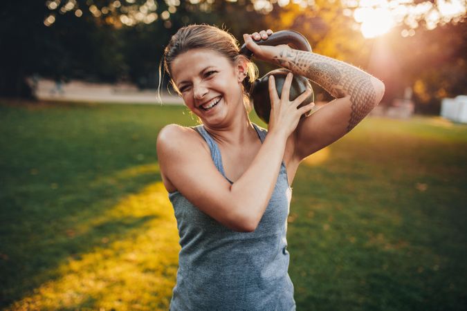 Portrait of happy young woman with kettlebell weights on her shoulder in the park