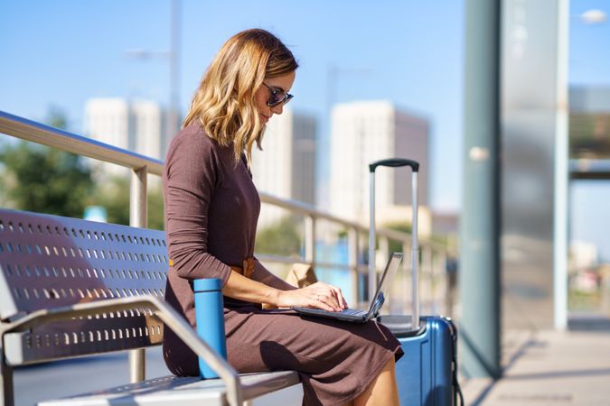 Side view of woman sitting on bench at station with laptop and suitcase