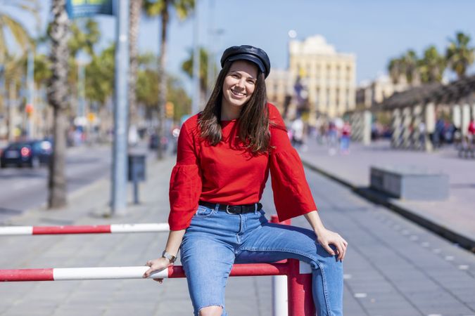Front view of a smiling beautiful young woman wearing red shirt sitting on a metallic fence and looking at camera outdoors in the street on a bright day