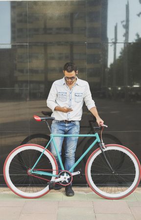 Male standing with his bike next to reflective wall looking down at phone