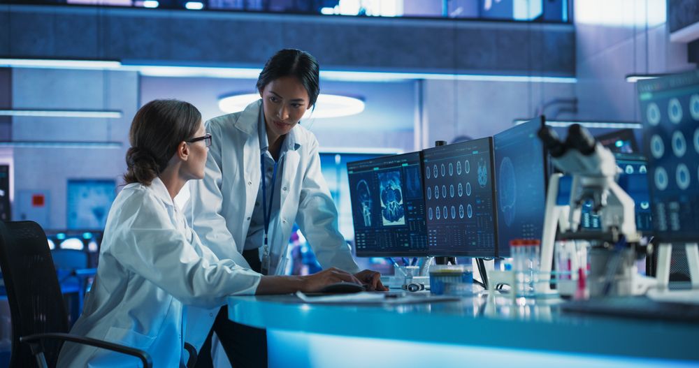 Asian And White Female Doctors Talking In Medical Research Center And Using Desktop Computer To Analyze MRI Scans Of Brain.