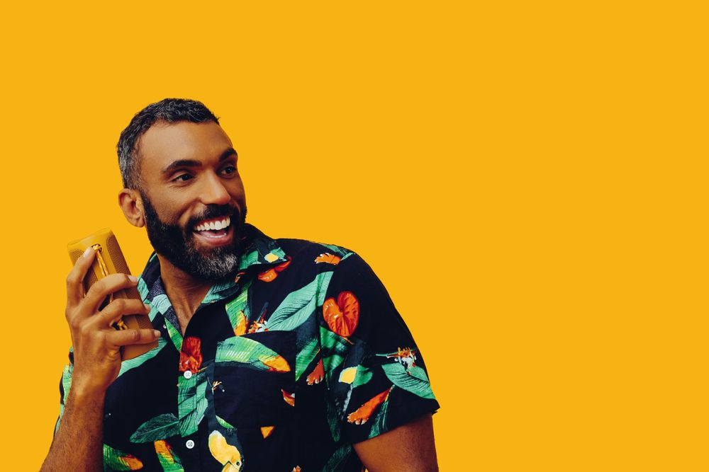 Black male in bold patterned shirt and small speaker up to his ear in yellow room