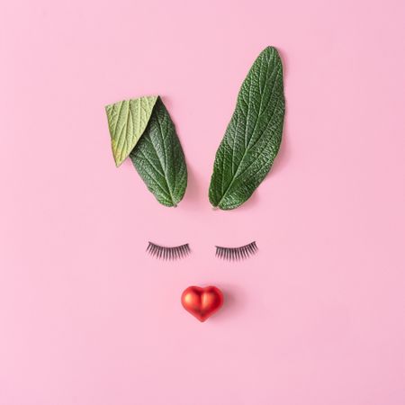 Bunny rabbit face made of green leaves with eyelashes and red heart on pastel pink background