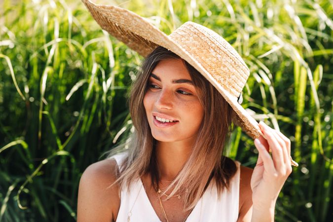 Woman sitting in front of long grass with hand on her straw hat