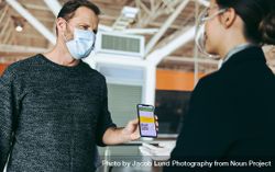 Male traveler in face mask showing his vaccine passport on smartphone to ground attendant 5pVX8b