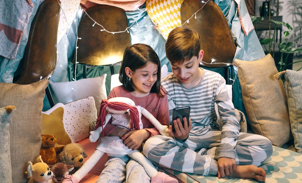 Happy children laughing together while watching video on cell phone sitting on shelter tent at home