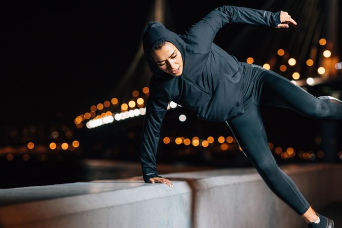 Woman in dark sportswear doing jumps on riverbank at night by lit bridge