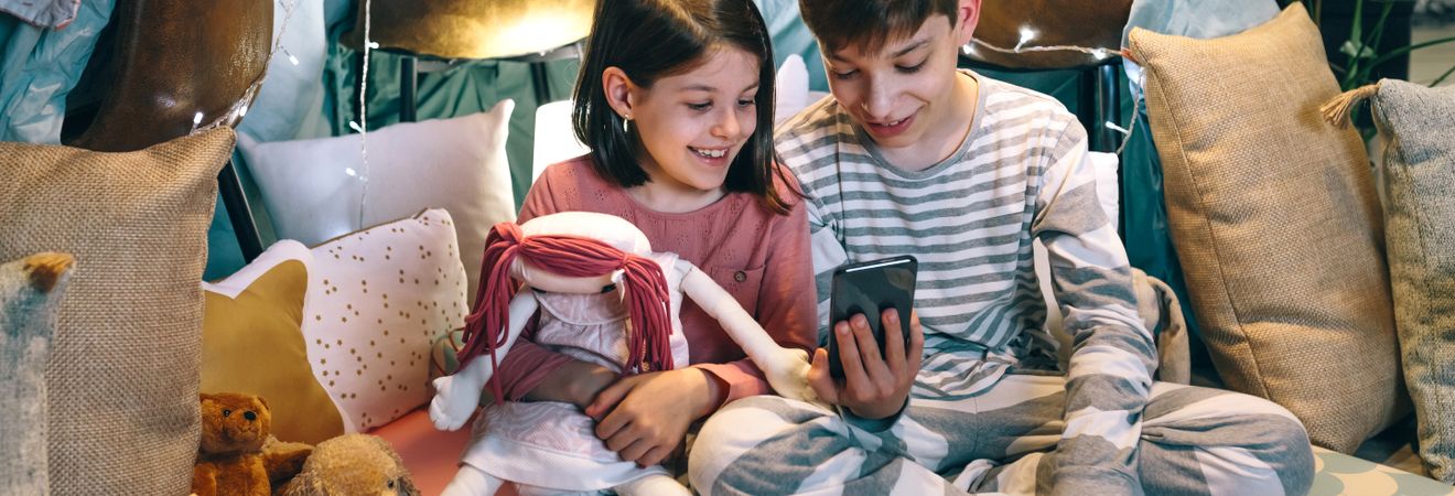 Banner of happy children laughing together while watch video on cell phone sitting on tent at home