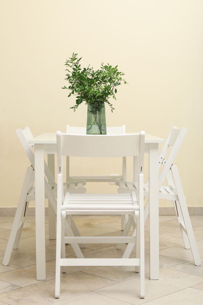 Wooden table with vase with plant and folding chairs against light beige wall