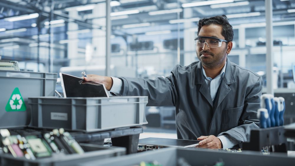 Indian Male Worker Disassembling Laptops To Recycle Electronic Components For Microchip Production At an Automated Electronics Factory With Robotic Arms.