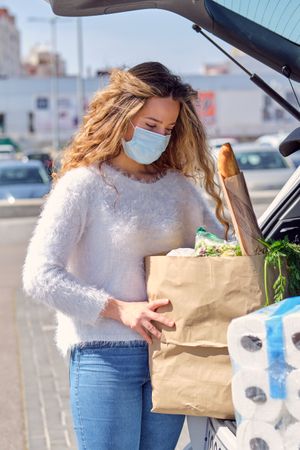 Woman in mask putting grocery bags in car trunk