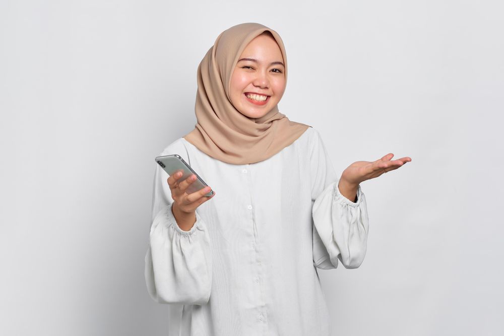 Asian female in headscarf smiling with one hand up and holding smart phone