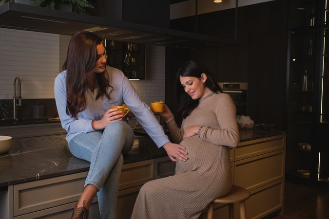 Expecting woman drinking tea with friend in kitchen