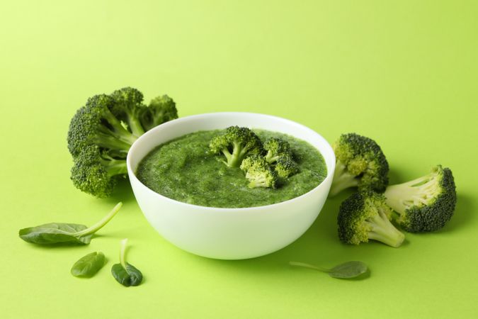 Bowl of broccoli soup surrounded with vegetables on green table