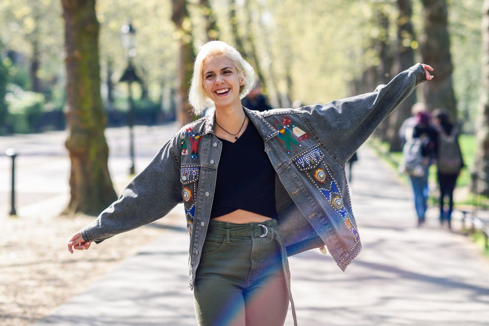 Blonde woman smiling in jean jacket with arms outstretched while walking in park