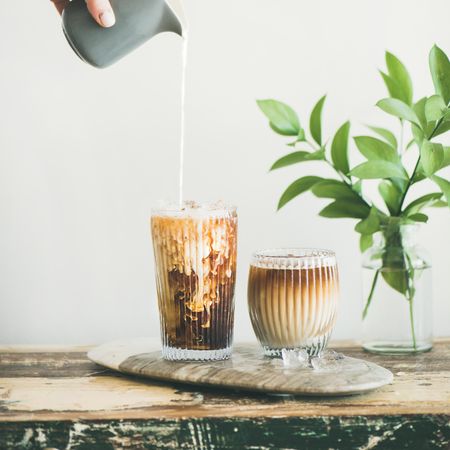 Hand pouring cream into iced coffee, square crop with leaves in background