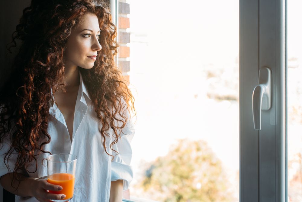 Woman standing by a windows blind drinking orange juice