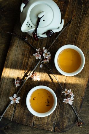 Top view of cups of green tea and pot with floral petals on wooden board