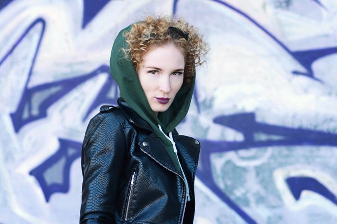 Fashionable portrait young woman hipster in green sweatshirt and leather biker jacket near the graffiti