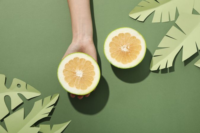 Two half of pomelo fruit, paper tropical leaves and hand on green background, top view