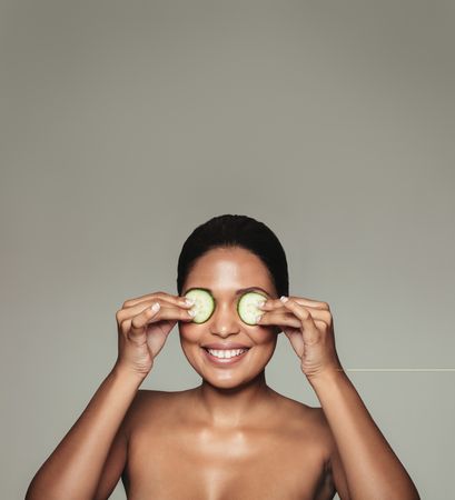Smiling female with clean skin holding cucumbers to her eyes