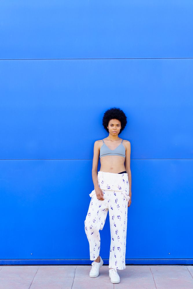 Woman in floral coveralls looking at camera while leaning against blue outdoor wall, copy space