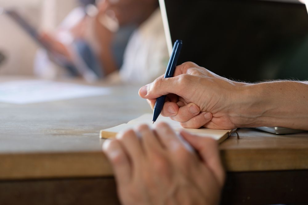 A close-up shot of a female hand holding a pen and writing in a notebook in an office setting