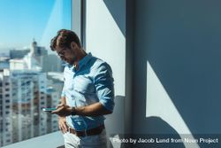 Professional male looking at cell phone next to glass window in office 5lG1v0