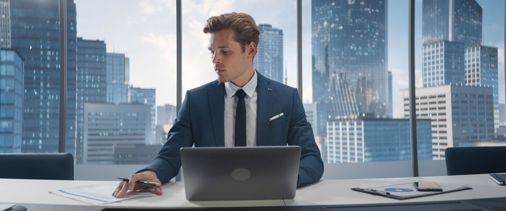 Portrait of Successful White Businessman Working on Laptop Computer in His Big City Corner Office.