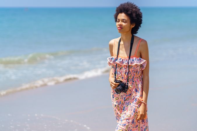 Confident female in maxi dress walking along coast with camera