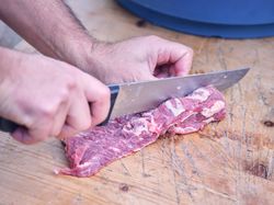Crop male cutting pork with knife on wooden table