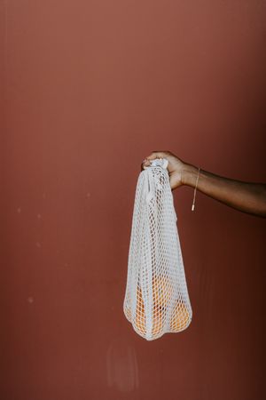 Person holding a mesh bag with oranges against red background