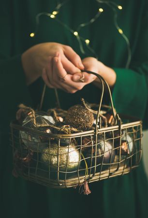 Woman in green dress holding wire basket of gold holiday decorations, vertical composition