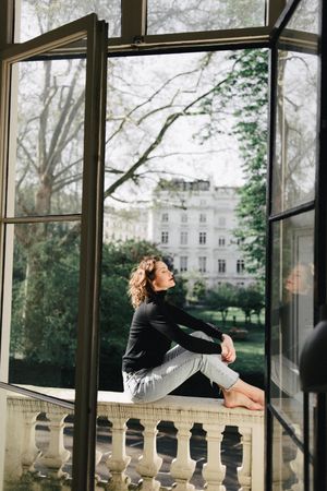 Side view of woman in casual outwear sitting on balustrade on balcony