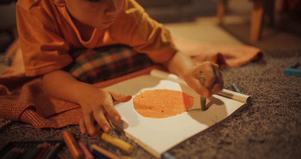 Close Up Shot Of Adorable Korean Boy Laying on The Floor And Drawing In Notebook With Pencils And Pens.