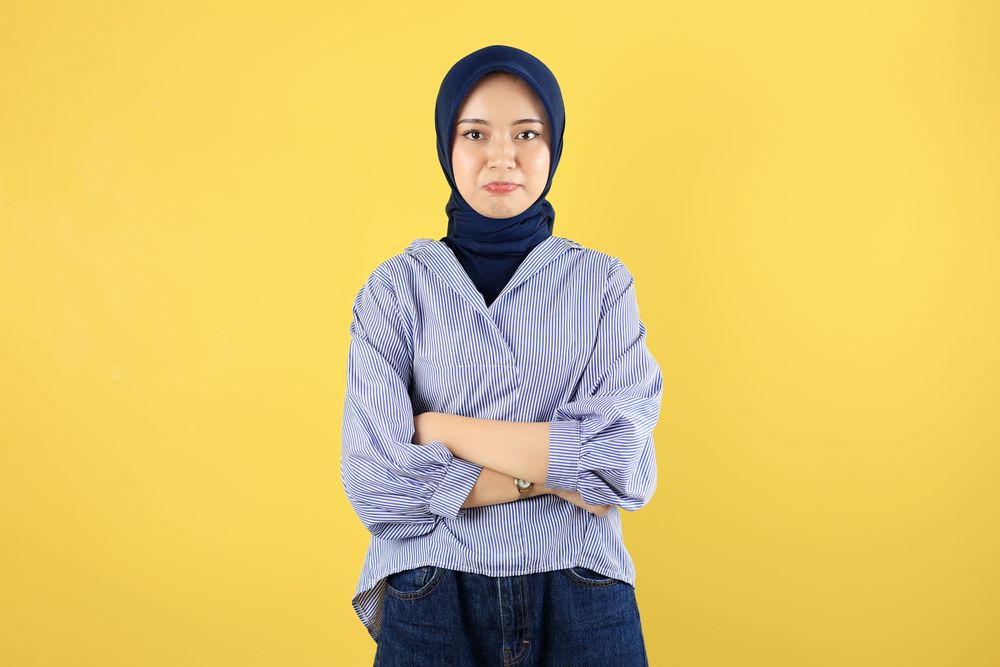 Displeased Young Asian Woman in Blue Striped Blouse and Navy Hijab Standing with Arms Crossed
