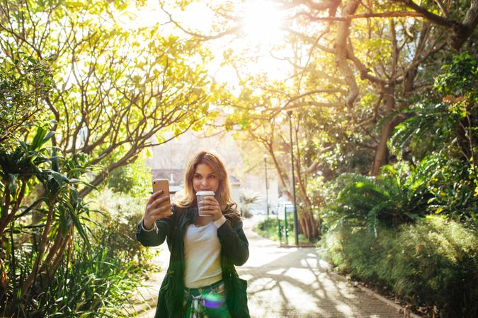 Woman taking a selfie in park holding a takeaway coffee