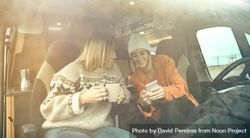 Female friends sitting in parked van having coffee and checking social media on phone 4dNDE0