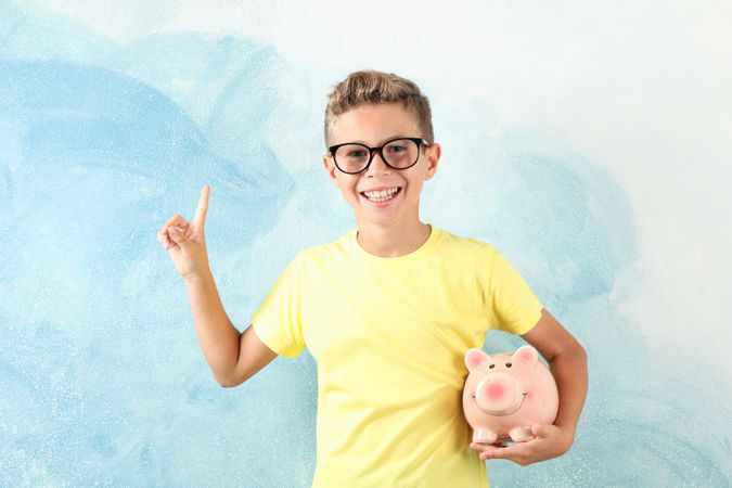 Boy in glasses with piggy bank against color background, space for text